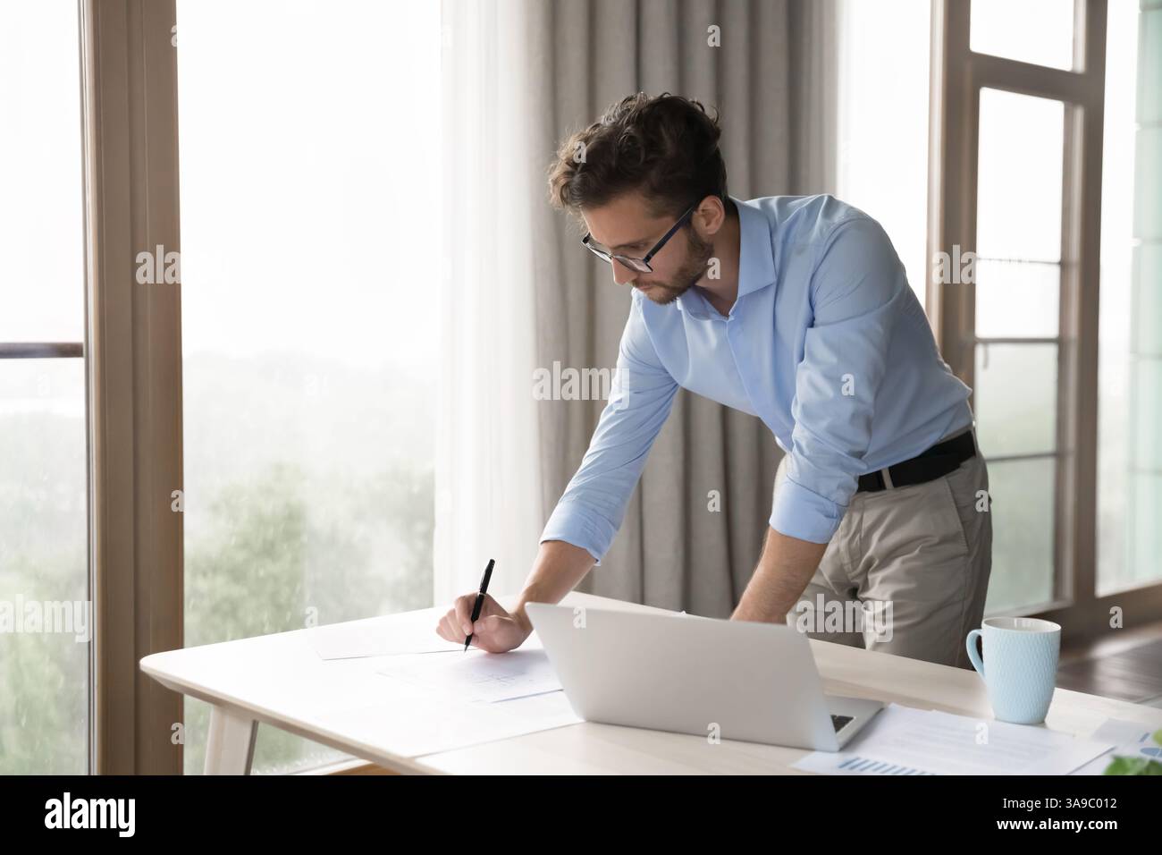 Young man standing by table review paper documents using laptop Stock Photo