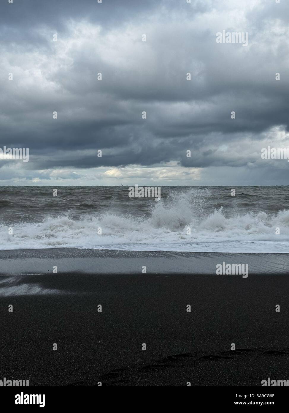 Rough ocean waves crash onto a black sand beach under a dark, cloudy ...