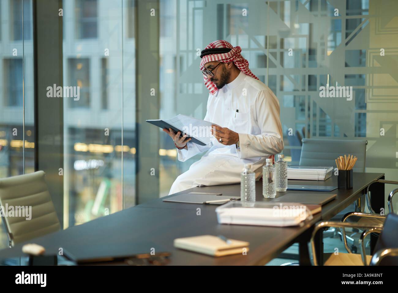 Serious young Middle Eastern businessman in traditional clothes sitting on table in board room working with important documents Stock Photo