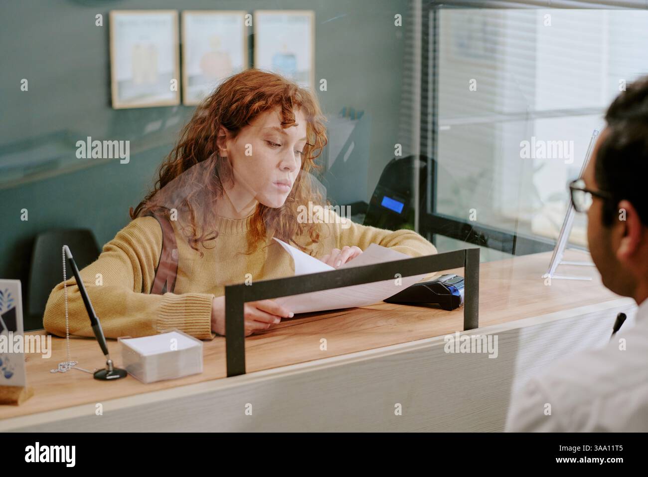 Young woman with curly hair sitting at reception desk while handling paperwork, interacting with unseen individual, including various documents and office supplies for transaction Stock Photo