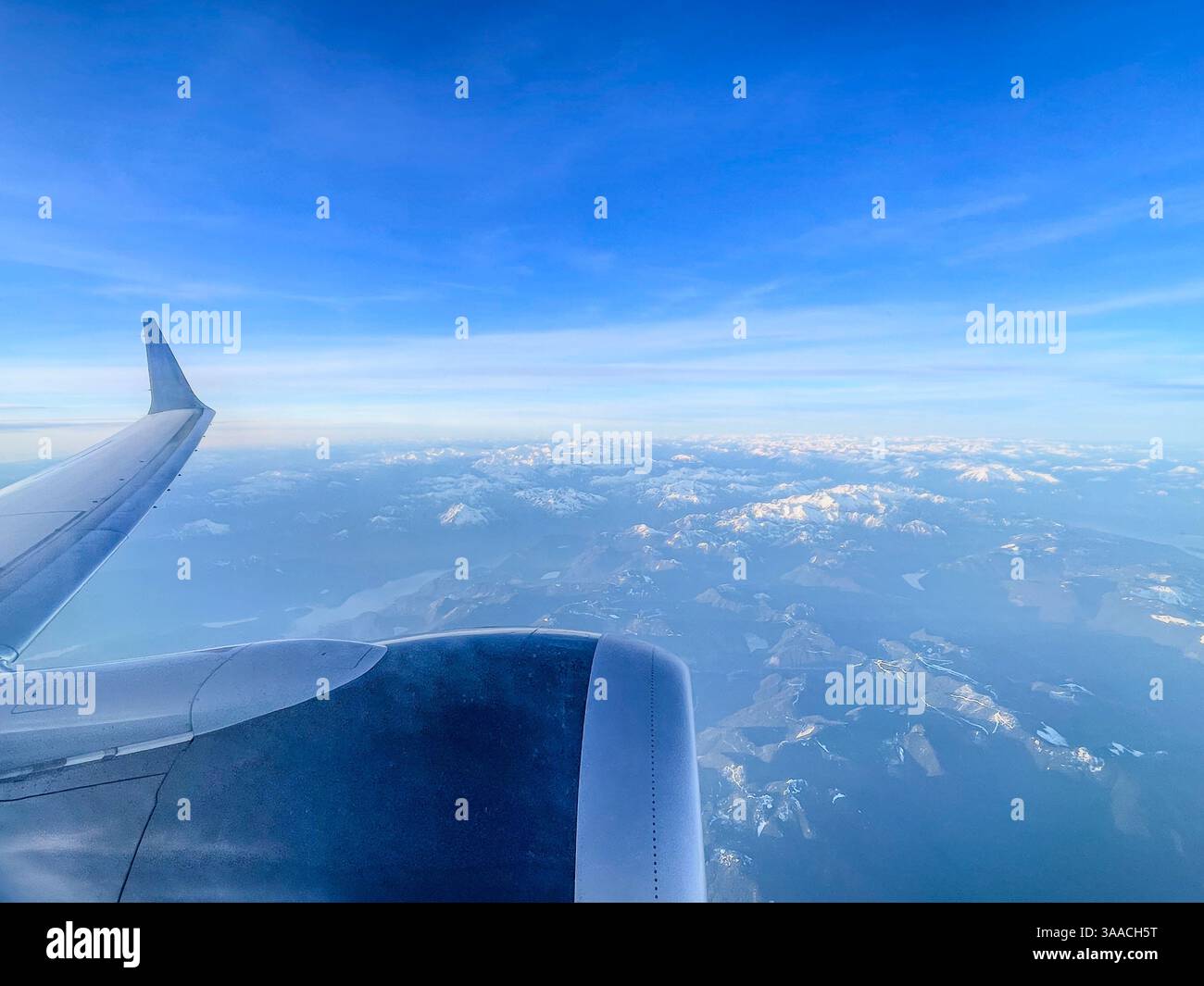 A view of an airplane wing flying over a snow-covered mountain landscape. Captured from high altitude with clear skies and mountain views. Stock Photo