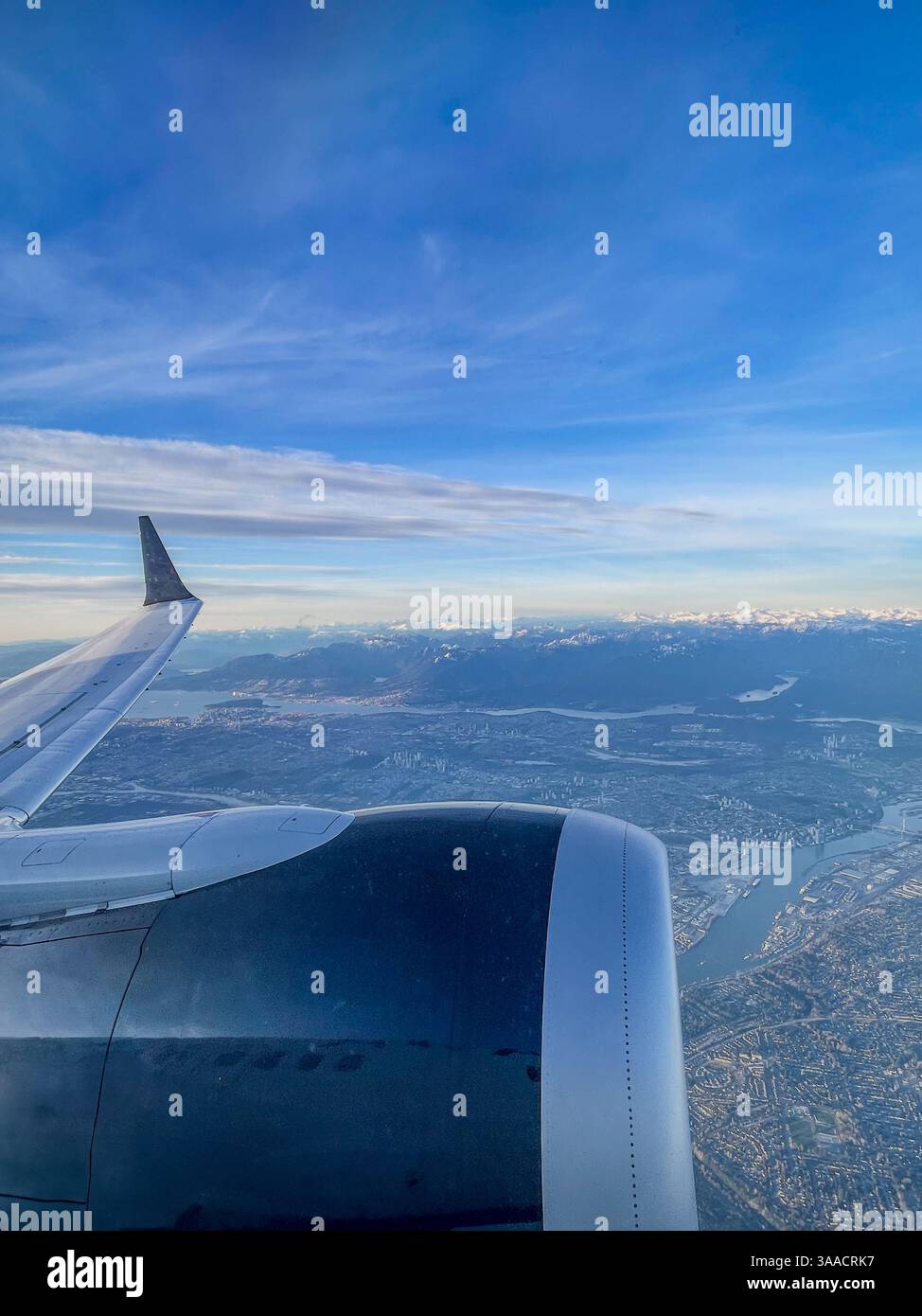 View of airplane wing and engine flying above a city with mountains and water in the background under a blue sky. - Smartphone Captured Stock Image