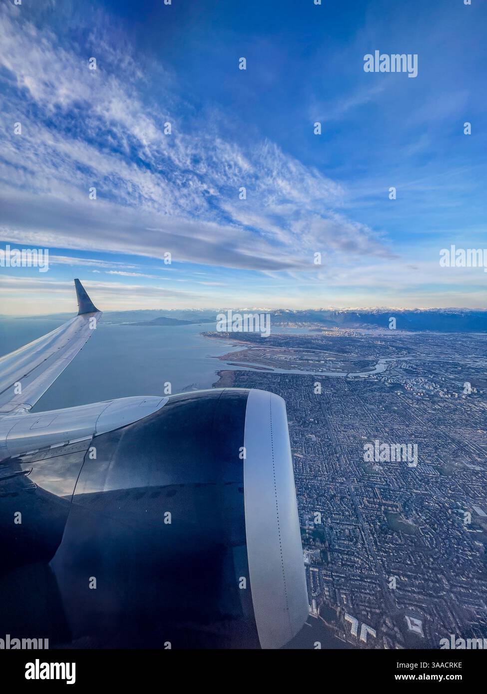 Airplane wing and engine view with a coastal city and mountains below. The image captures a high-altitude perspective during daytime. - Smartphone Captured Stock Image