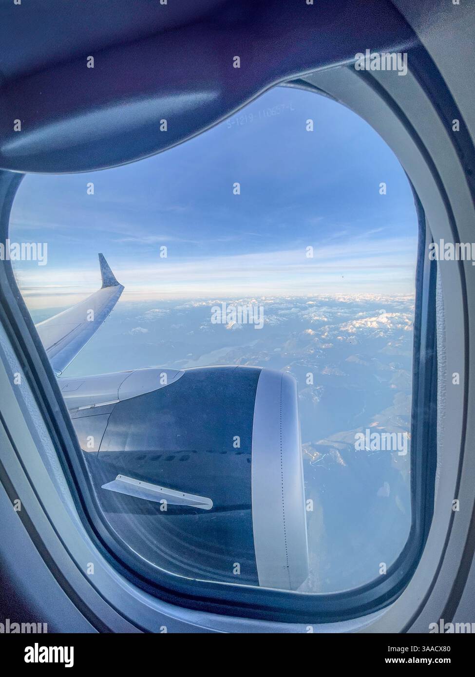 View of an airplane wing and engine through a passenger window, capturing snowy mountain peaks in the background during high-altitude travel. - Smartphone Captured Stock Image