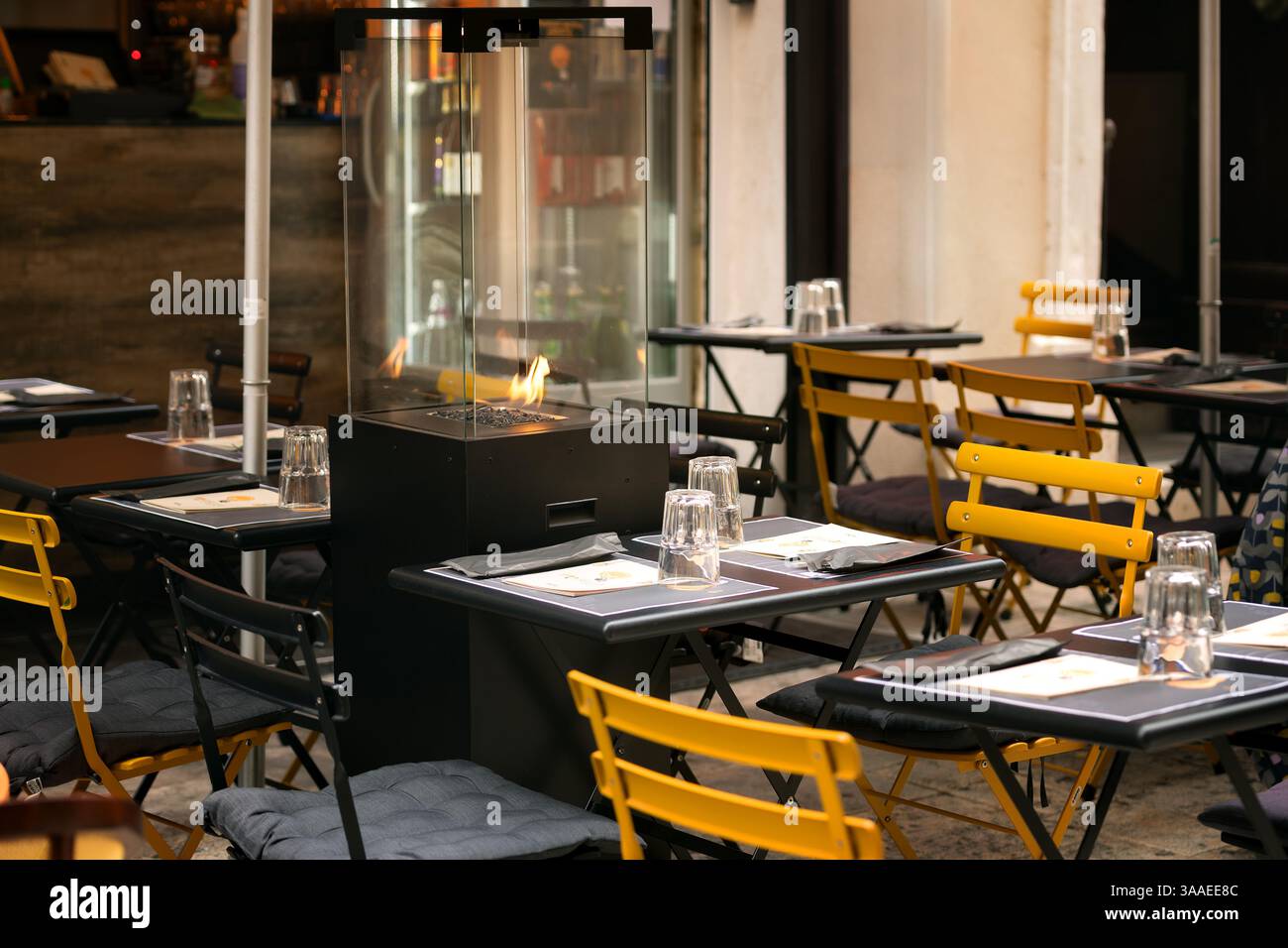 Outdoor restaurant seating with black and yellow chairs, set tables, and a modern glass-enclosed fireplace. Cozy evening dining ambiance in a European Stock Photo