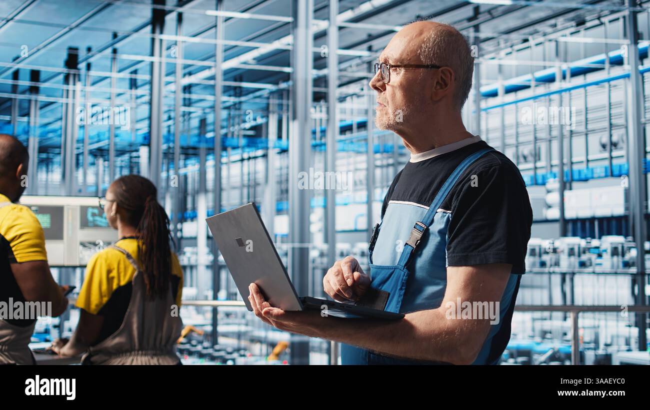 Smart factory senior engineer uses laptop to oversee operation of robotic arms and conveyor systems. Experienced industry 4.0 plant technician monitors system performance using notebook, camera A Stock Photo