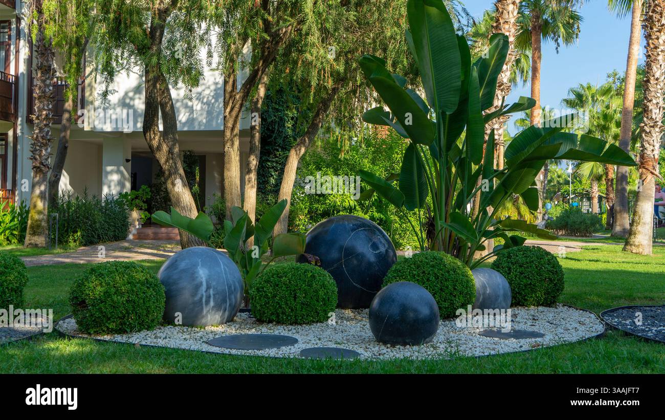 Mediterranean style landscaping, banana palm and bushes in form of balls and gray and black stone balls on small light pebbles and green lawn. Stock Photo