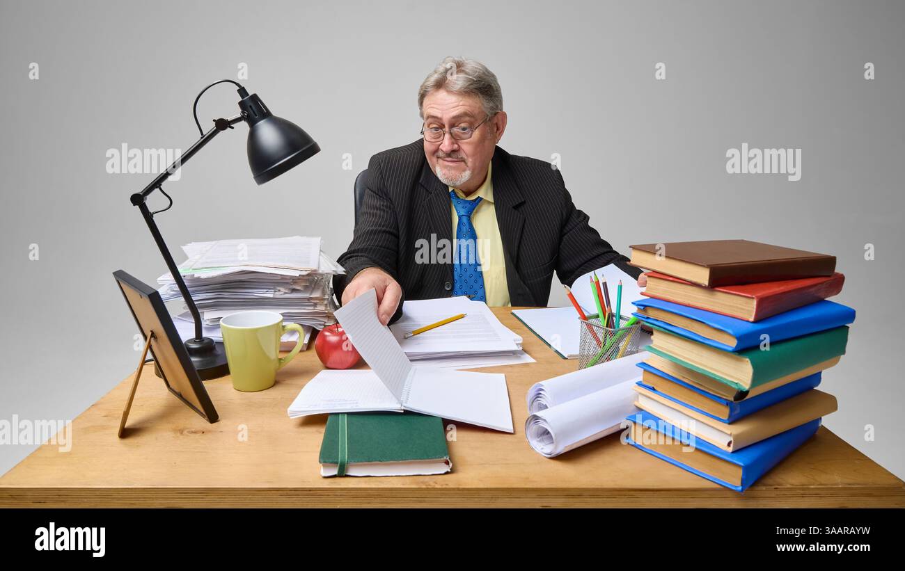 Senior man checking documents at cluttered desk with skeptical expression against light grey background. Stock Photo