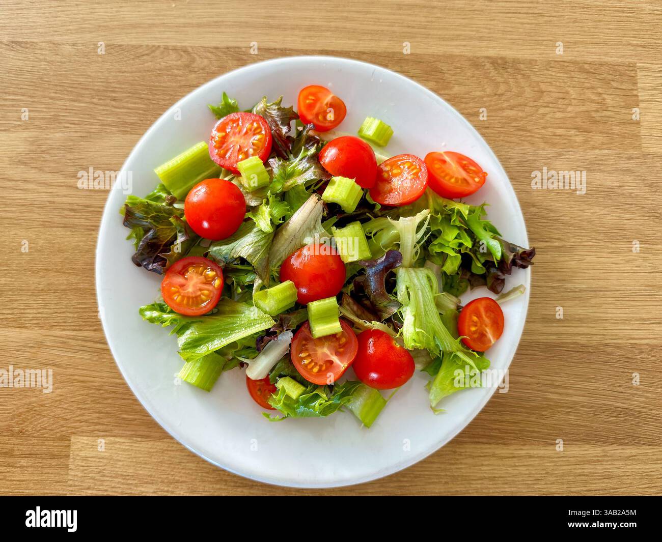 Simple salad of tomatoes, celery, and mixed lettuce leaves on a small white plate on a wooden table. No people. Diet concept. - Smartphone Captured Stock Image