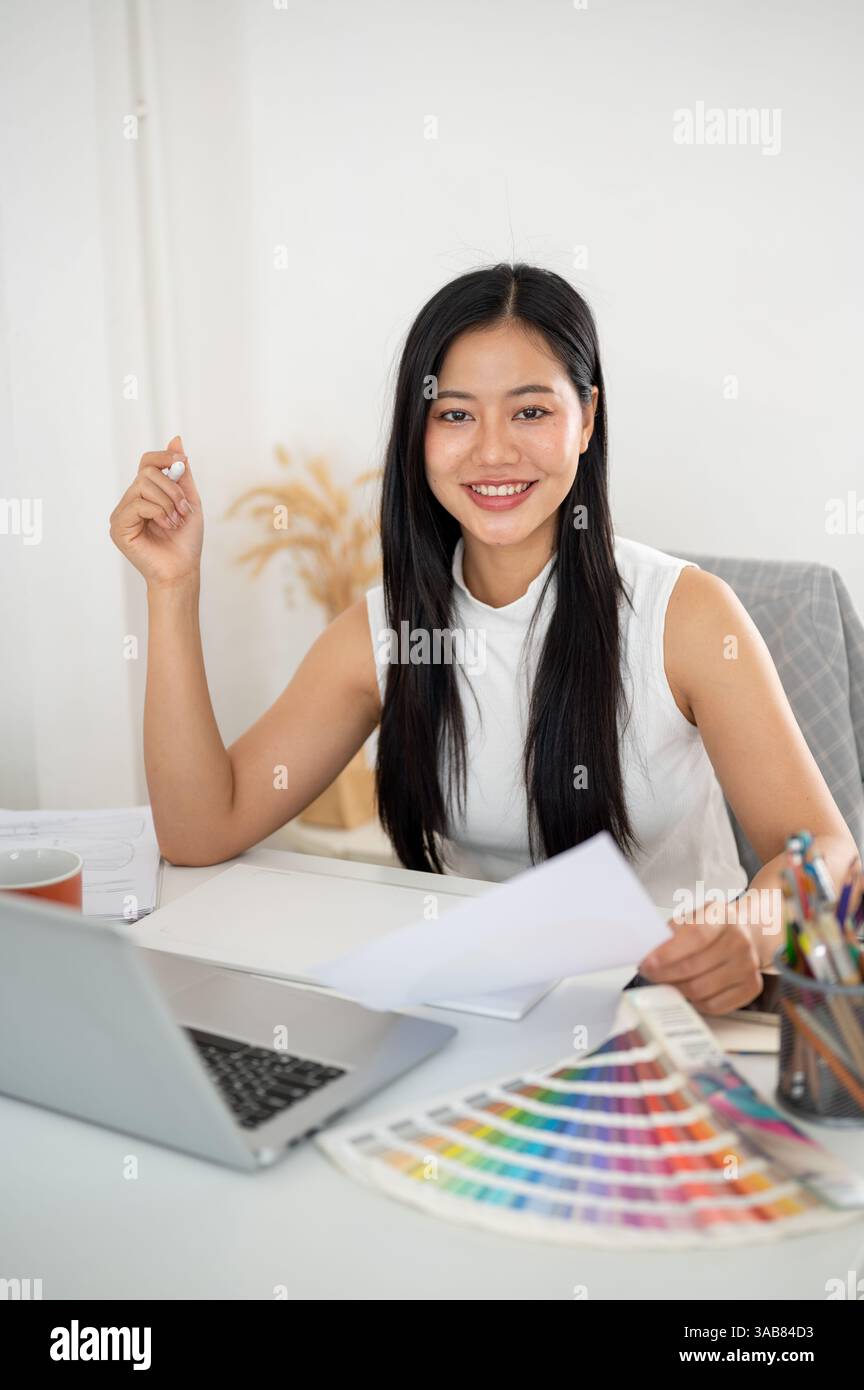 An asian woman is smiling at camera while holding the documents about design color on her working desk. Interior Designer or Graphic Designer working Stock Photo