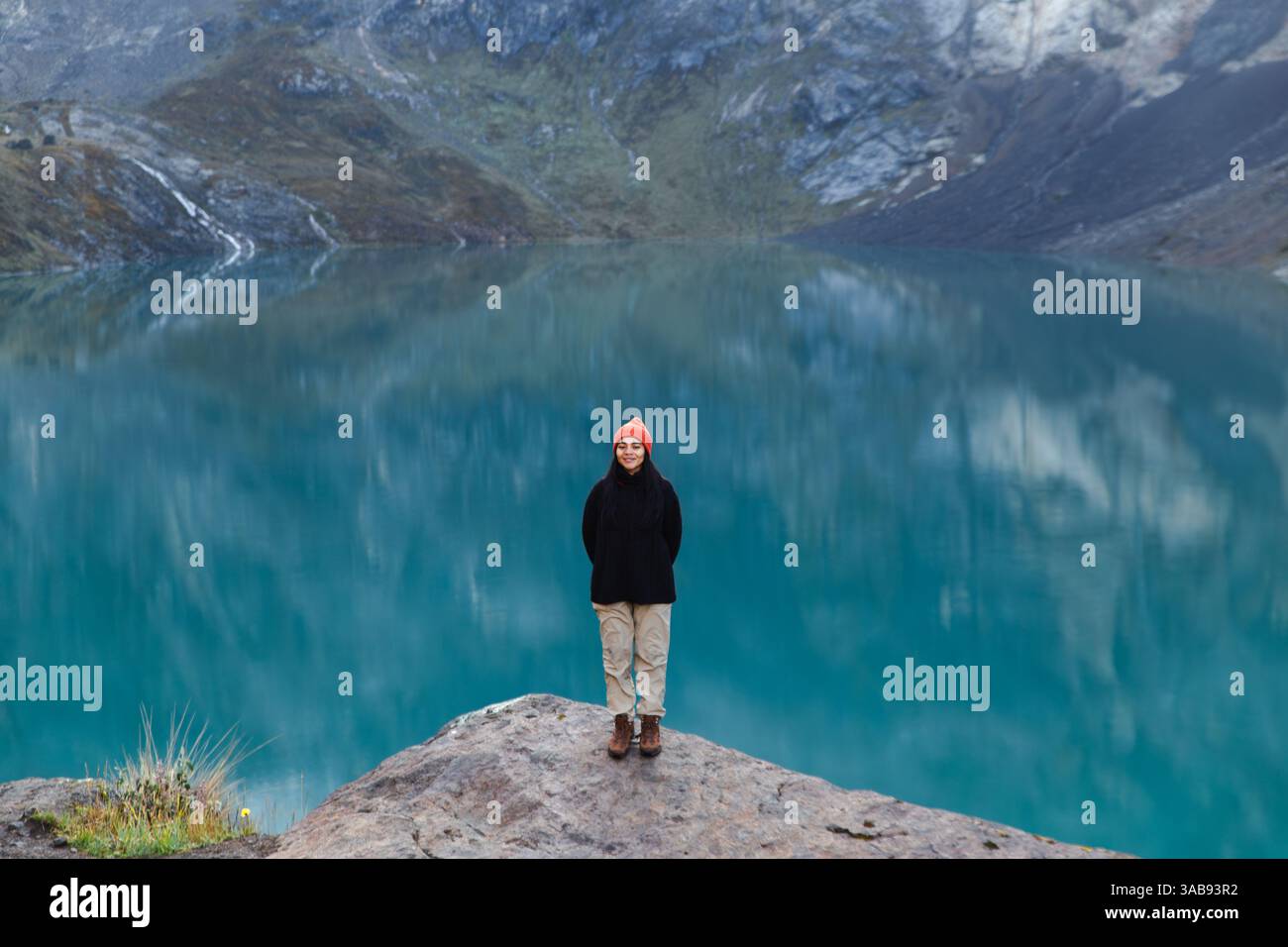 An Ecuadorian woman stands on a rock by the turquoise lagoon at El Altar volcano, Ecuador. The breathtaking landscape features serene water and rugged Stock Photo