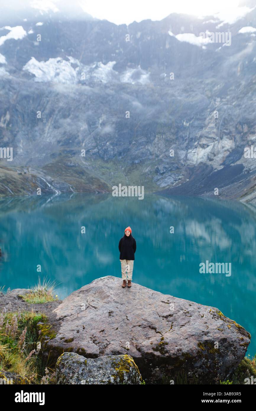 An Ecuadorian woman in a hat and jacket stands on a rock overlooking the turquoise lake at El Altar Volcano, Ecuador, with majestic mountains in the b Stock Photo
