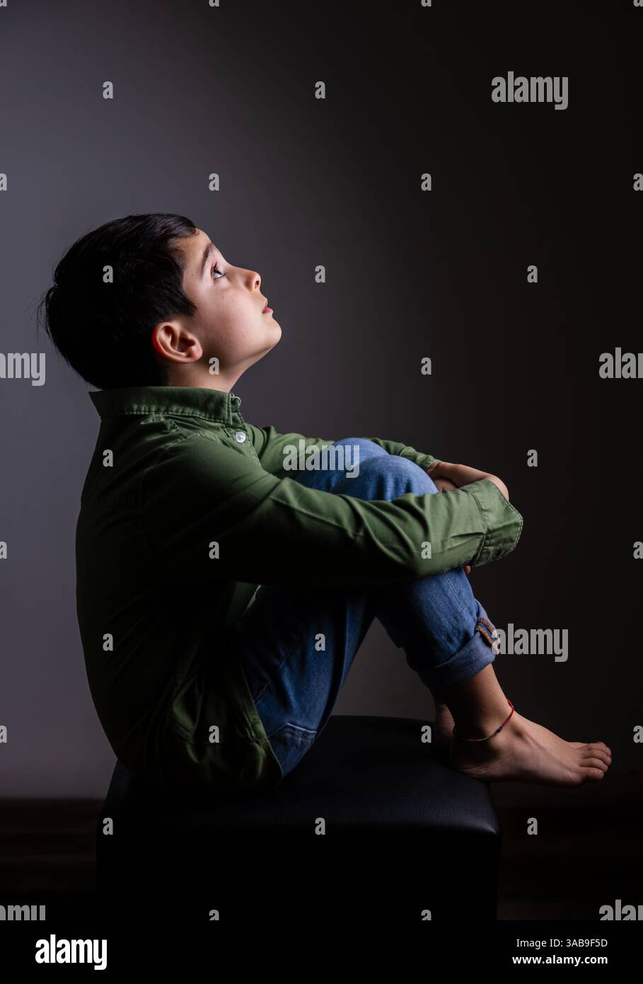 Young boy sits on a stool in a dark room, looking upward thoughtfully ...