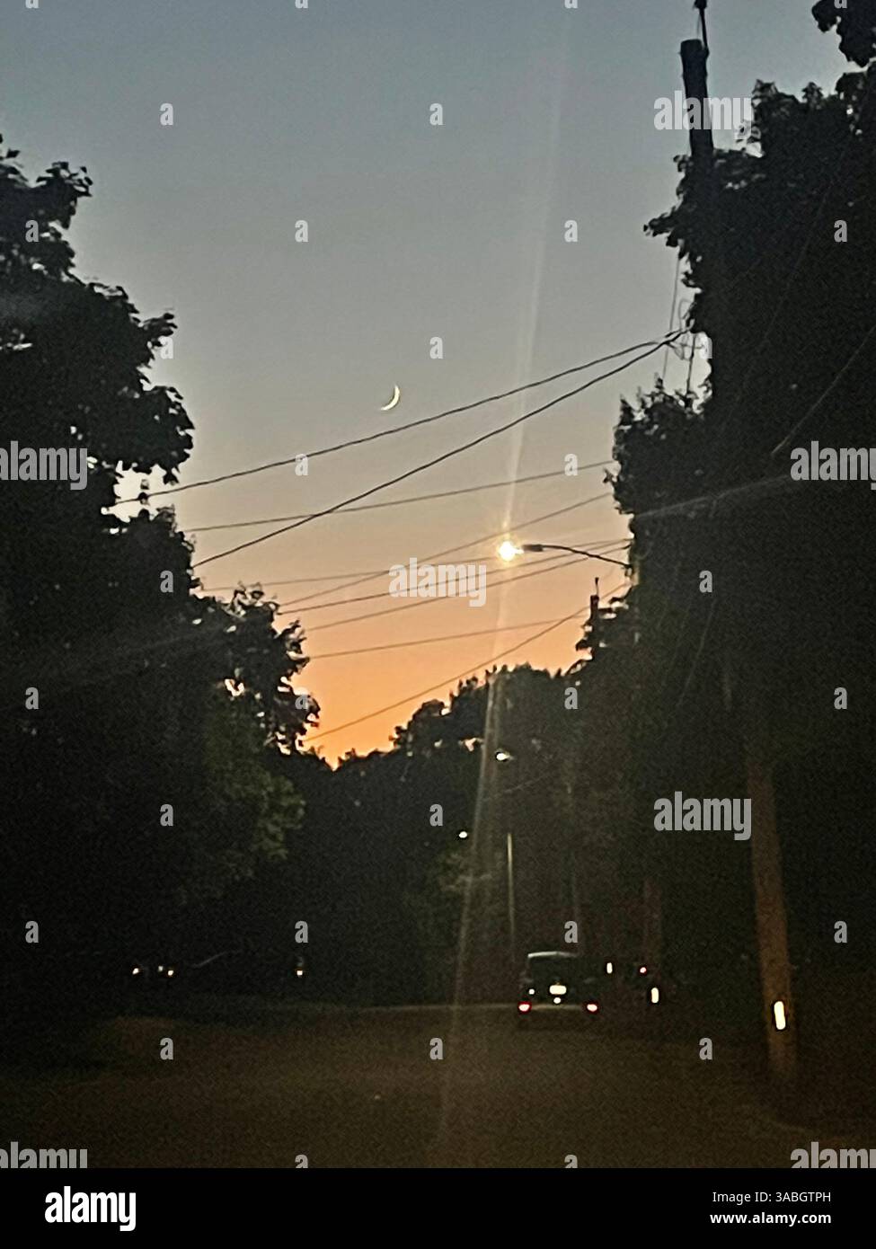 A peaceful suburban street at dusk with a glowing streetlight and a crescent moon in the sky. The scene captures the transition from day to night. - Smartphone Captured Stock Image