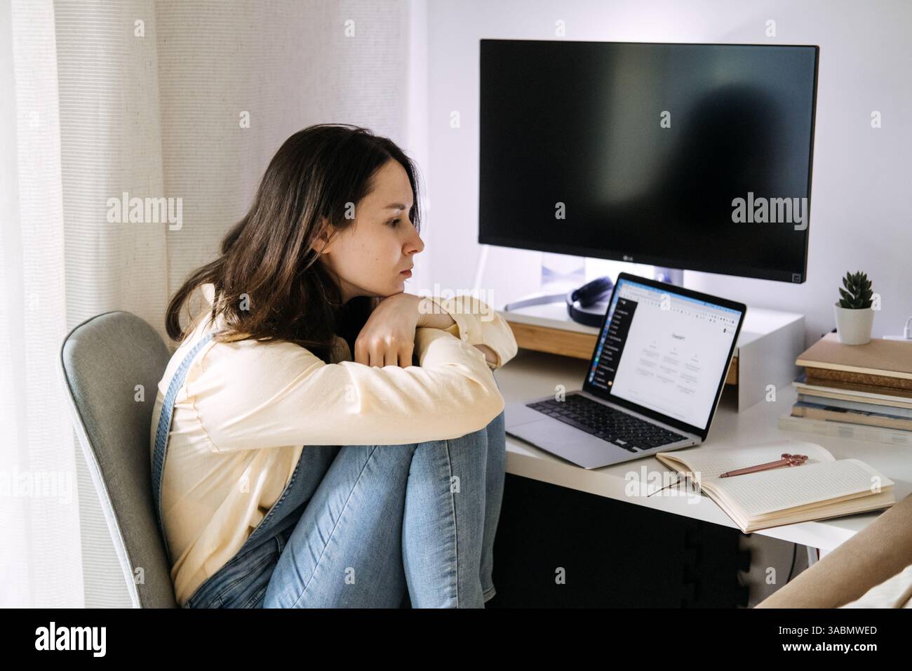 Young Asian woman sitting curled up at her desk, looking at a laptop displaying a ChatGPT interface. Creativity with ChatGPT, digital collaboration Stock Photo