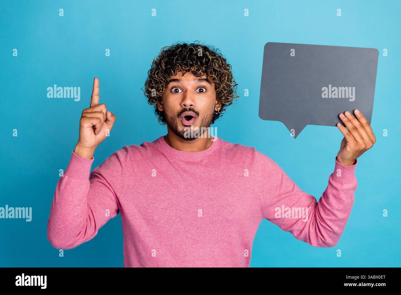 Surprised Young Man Holding Speech Bubble While Pointing Upwards Against Blue Background Stock Photo