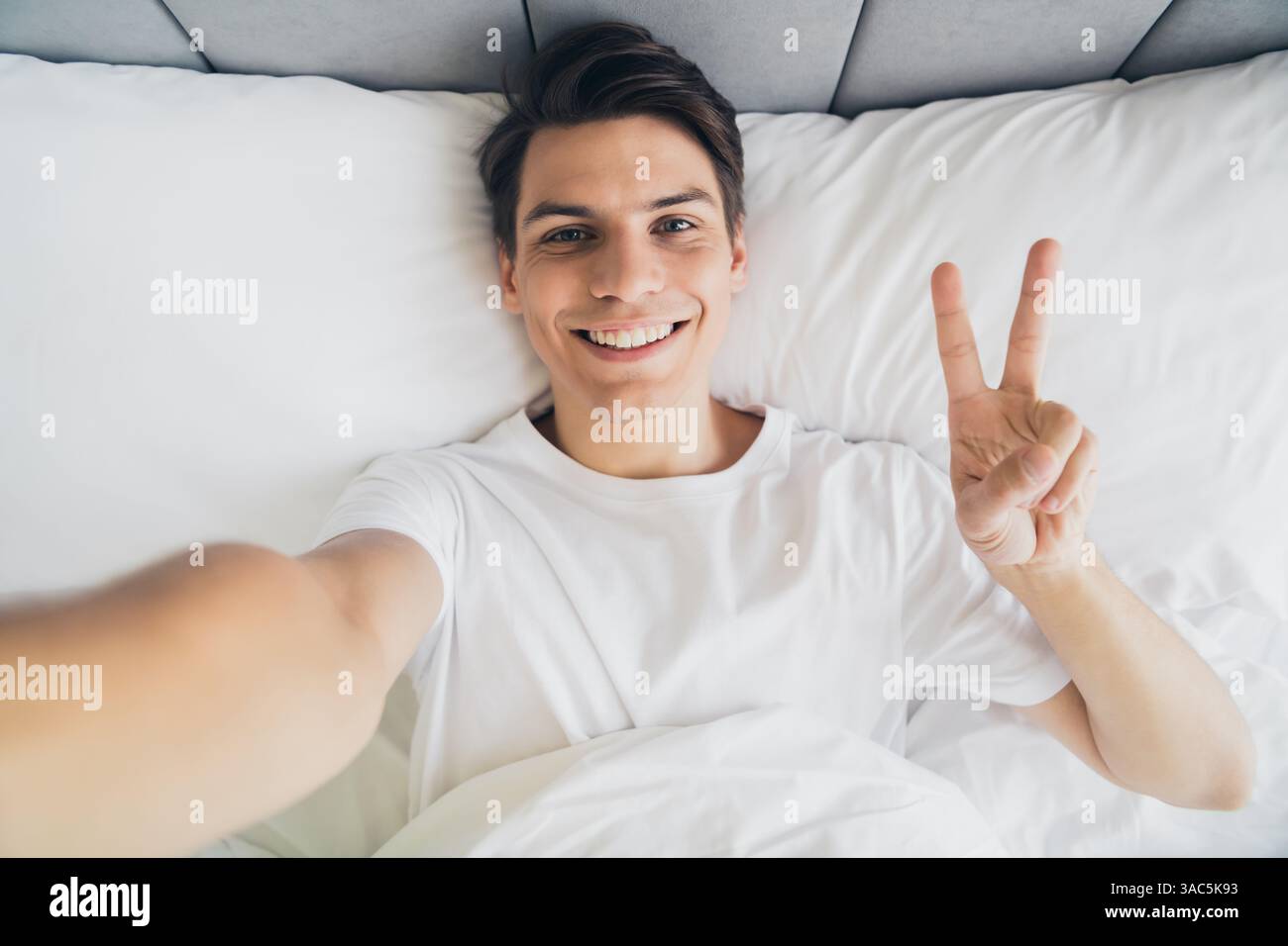 Bright young man in pajama relaxing on bed showing peace sign while smiling Stock Photo