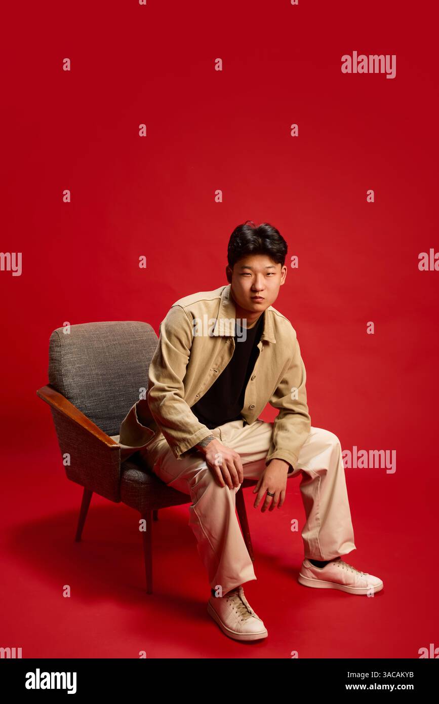 Thoughtful young man sitting in armchair leaning forward slightly with focused look against red backdrop. Concept of contemplation, self-expression Stock Photo