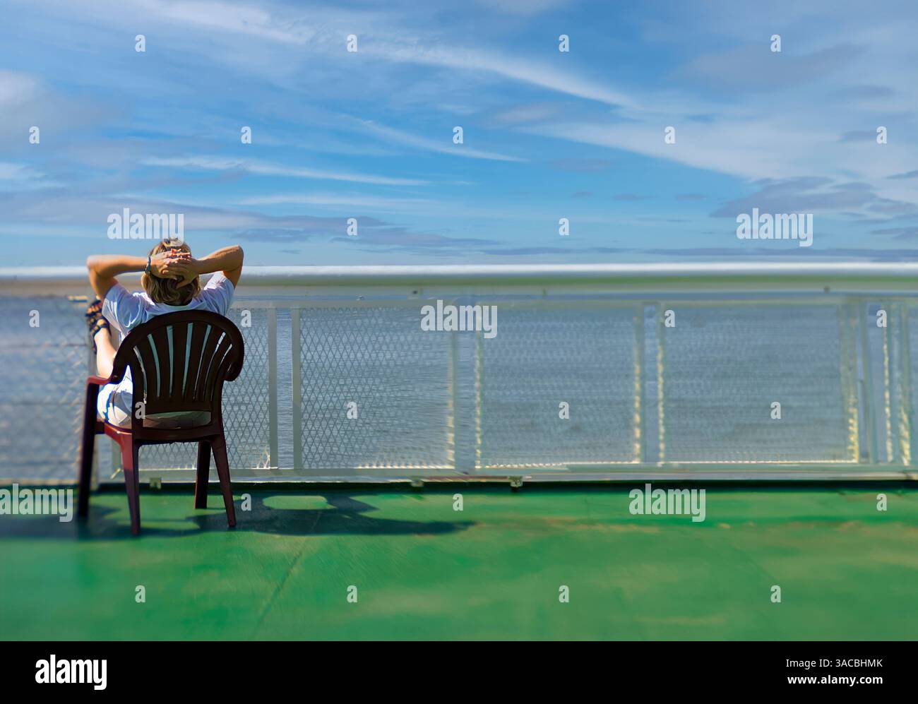 A person relaxes on a chair on the deck of a ferry, gazing out at a clear, tranquil water view under a blue sky with scattered clouds. Stock Photo