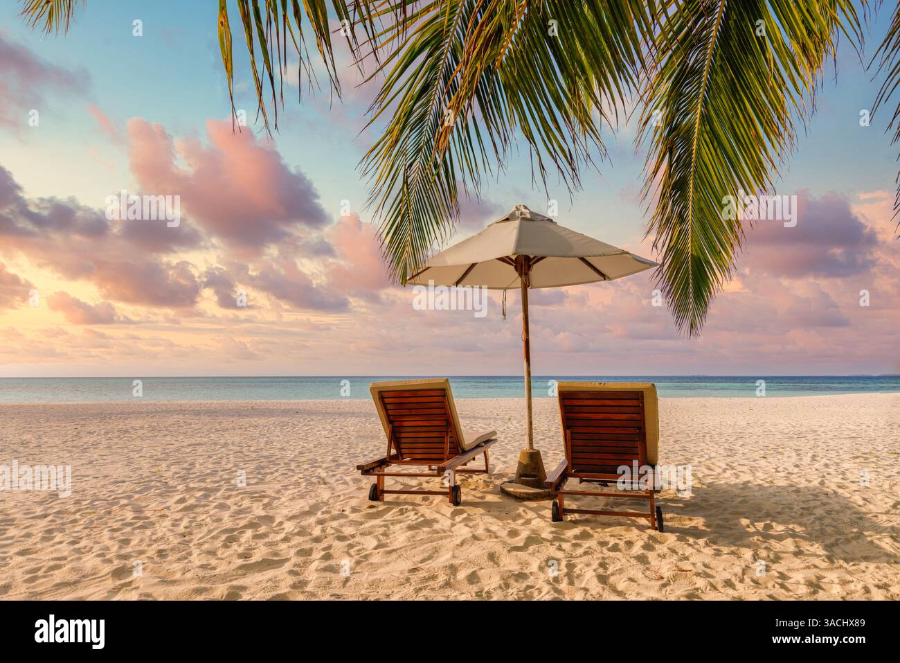 Sunset views tropical beach chairs umbrella on golden sand, serene turquoise sea, palm trees under vibrant colorful sky. Romantic honeymoon landscape Stock Photo