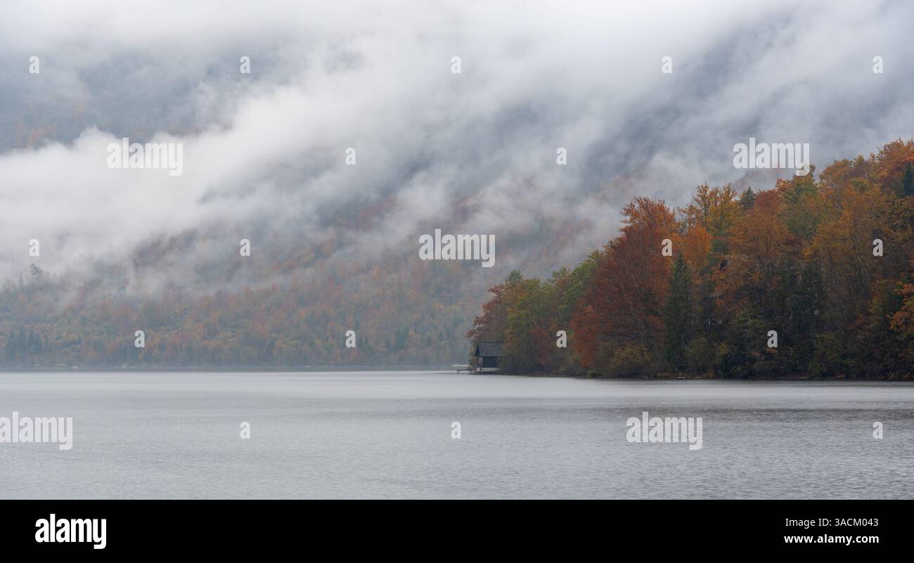Fog is rising from lake bohinj and covering the mountains in the background, creating a mysterious atmosphere during the autumn season Stock Photo