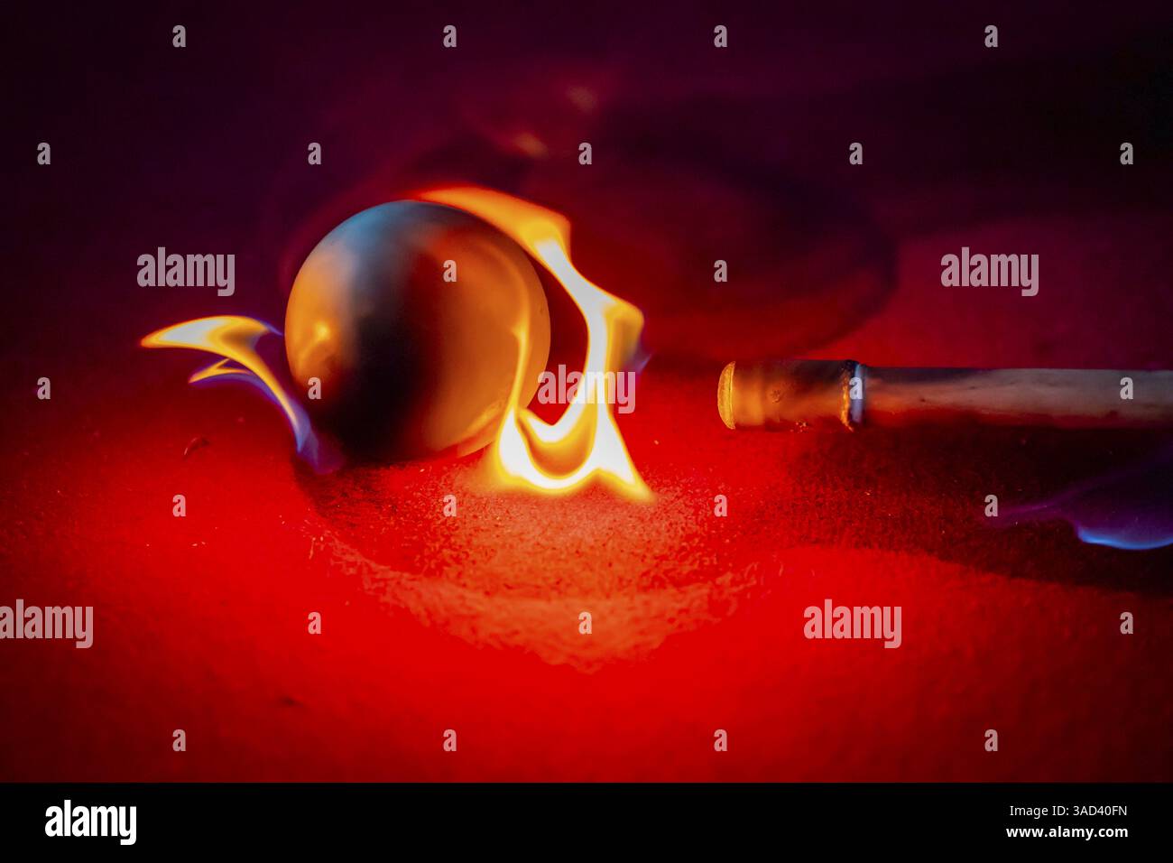 A billiard table is engulfed in flames, with blazing balls scattering across the charred red felt. Smoke curls upward, casting eerie shadows, as each Stock Photo