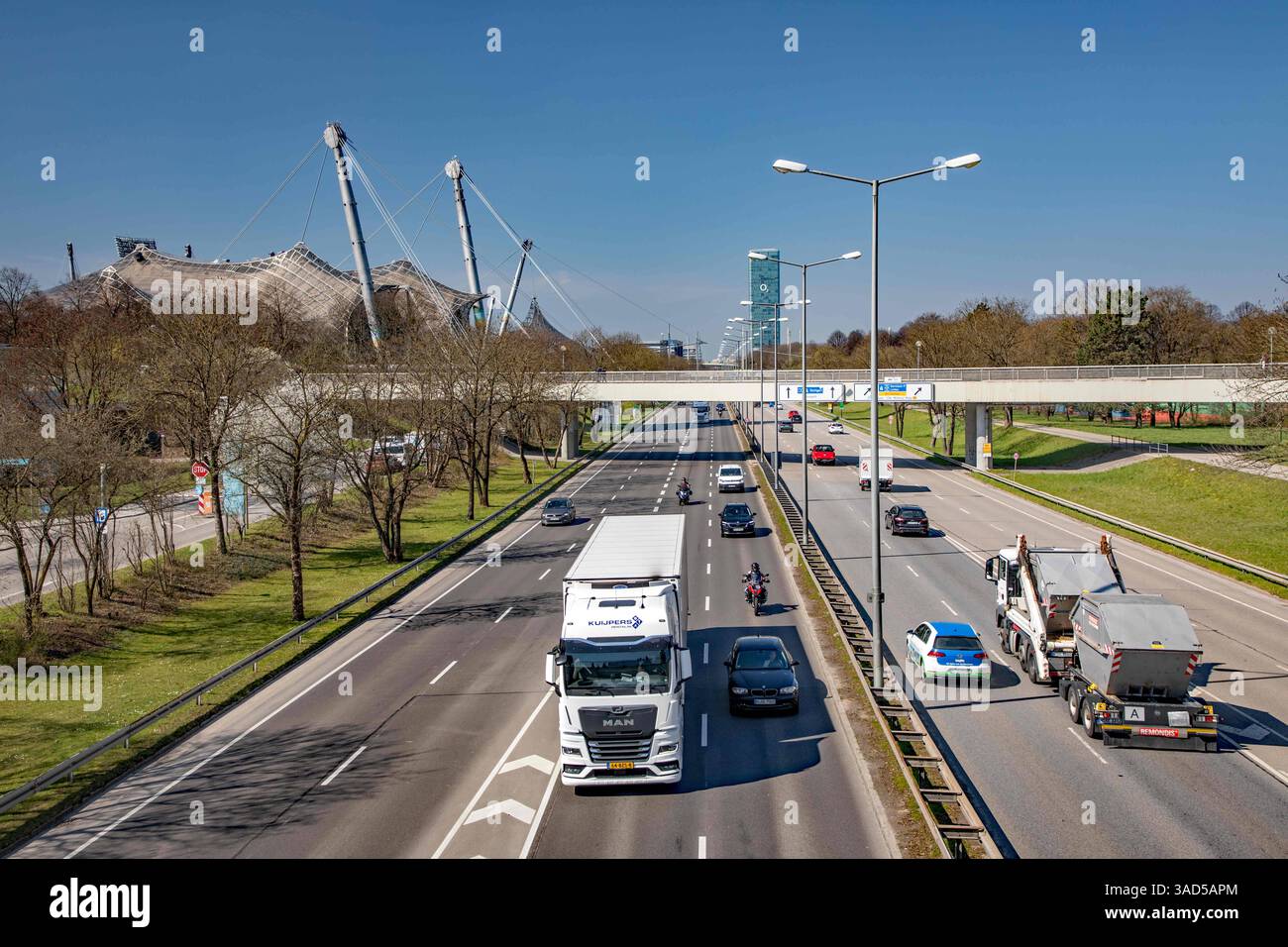 München, Deutschland 03. April 2025: Im Bild: Blick auf den Mittleren Ring im Olympiapark, LKW und Container-Transporter, links ist das Zeltdach des Olympiastadions zu sehen, im Hintergrund der O2-Tower. Bayern *** Munich, Germany 03 April 2025 In the picture view of the Mittlerer Ring in the Olympic Park, trucks and container transporters, the tent roof of the Olympic Stadium can be seen on the left, in the background the O2 Tower Bavaria Copyright: xFotostandx/xFritschx Stock Photo