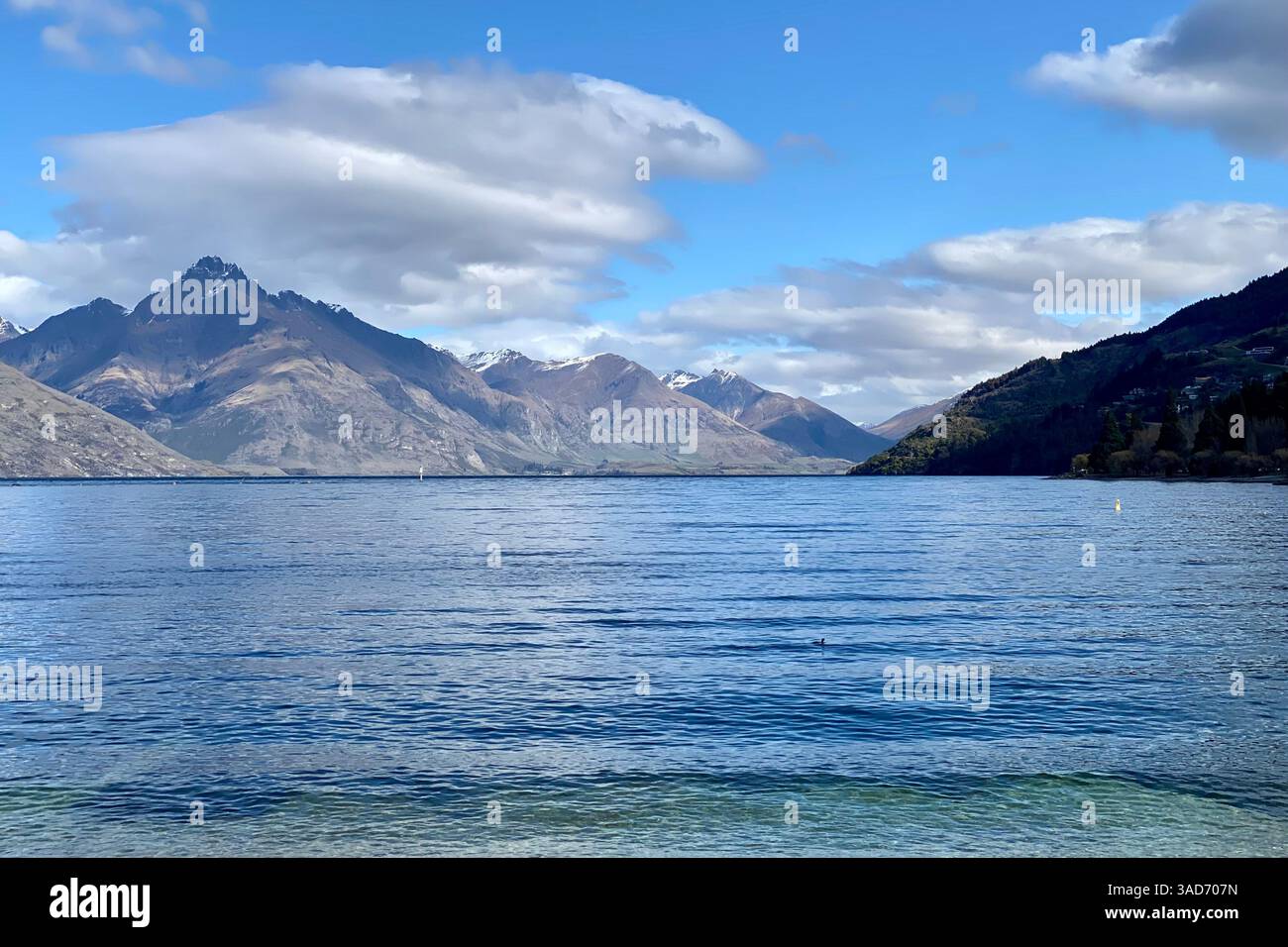 Crystal-clear Lake Wakatipu, Queenstown, reflects sunlit mountains and drifting clouds beneath a brilliant blue sky. Serene and majestic view. Stock Photo