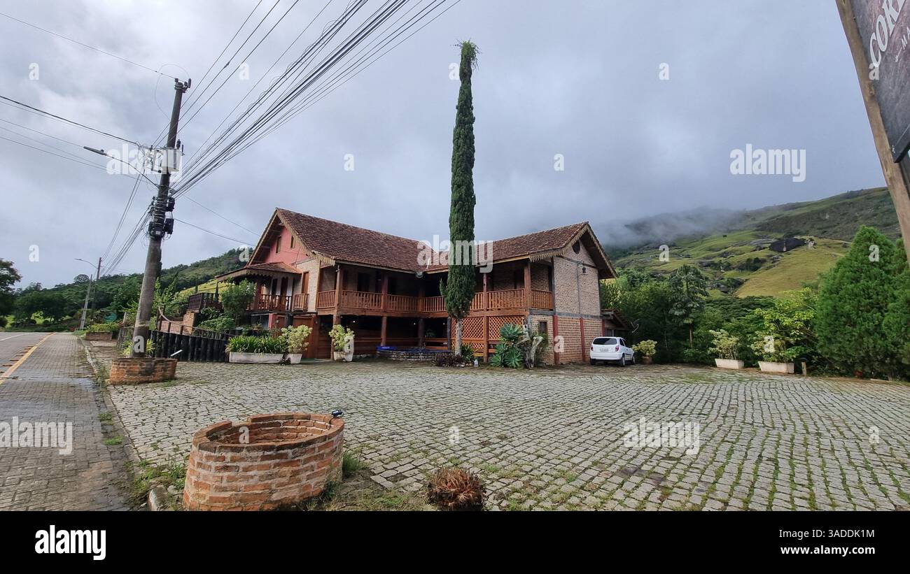 A picturesque countryside scene featuring a charming two-story brick house with wooden balconies and a tiled roof, set against a lush green mountain. - Smartphone Captured Stock Image