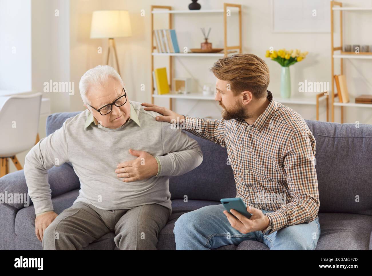 Worried man calling emergency ambulance to senior father suffering from heart attack at home. Stock Photo