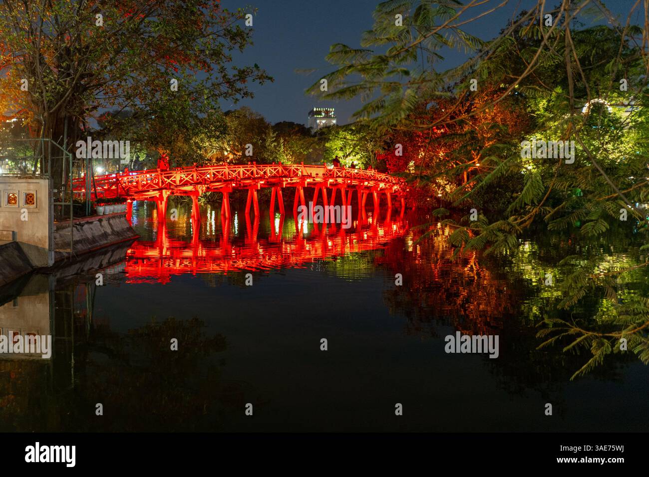 A vivid night scene captures the iconic The Huc Bridge in Hanoi, Vietnam, reflecting in the tranquil waters. The red bridge glows under the lights. Stock Photo