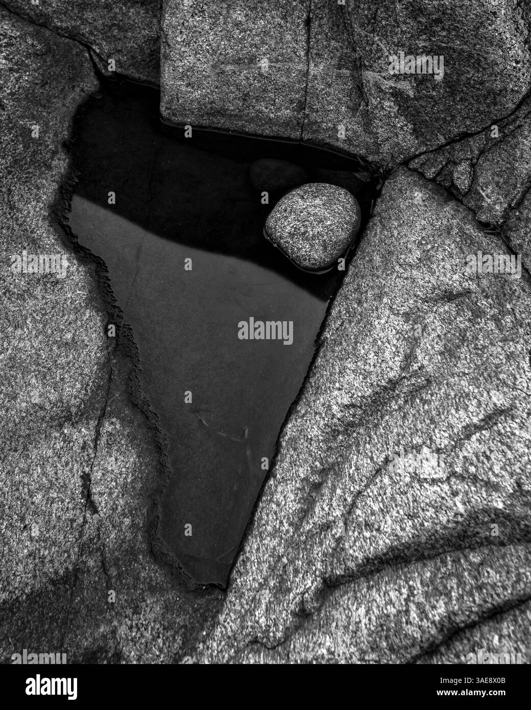Black and white depiction of coastal rocks in Sweden showcases unique formations and reflections in a small tide pool. The rugged shoreline creates a Stock Photo