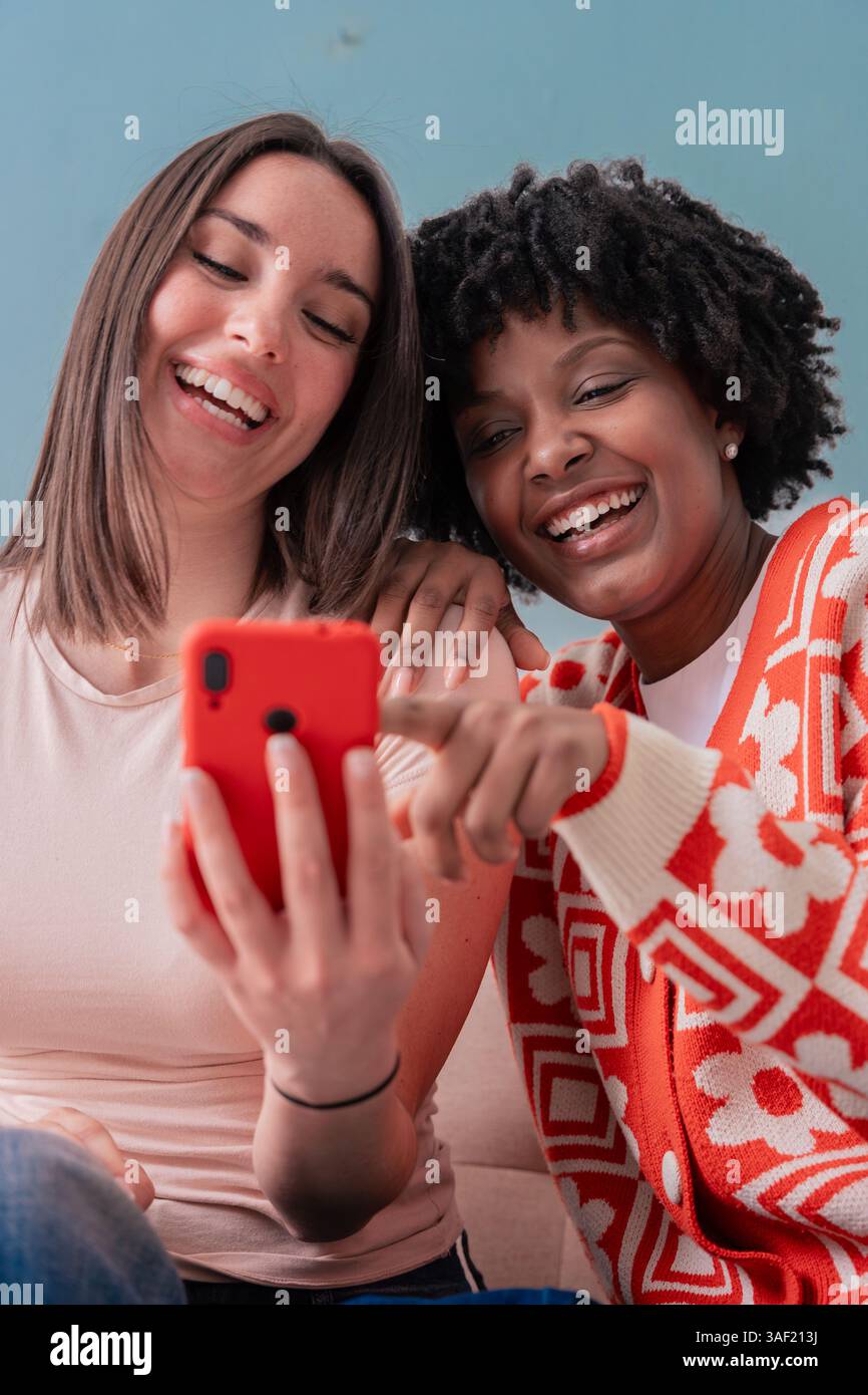 Two happy women sitting on couch smiling while looking at smartphone screen Stock Photo