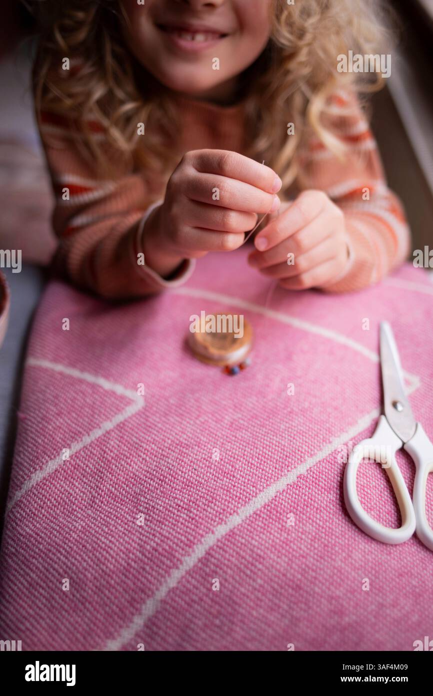 Child threading a needle while crafting on a pink textile surface Stock Photo