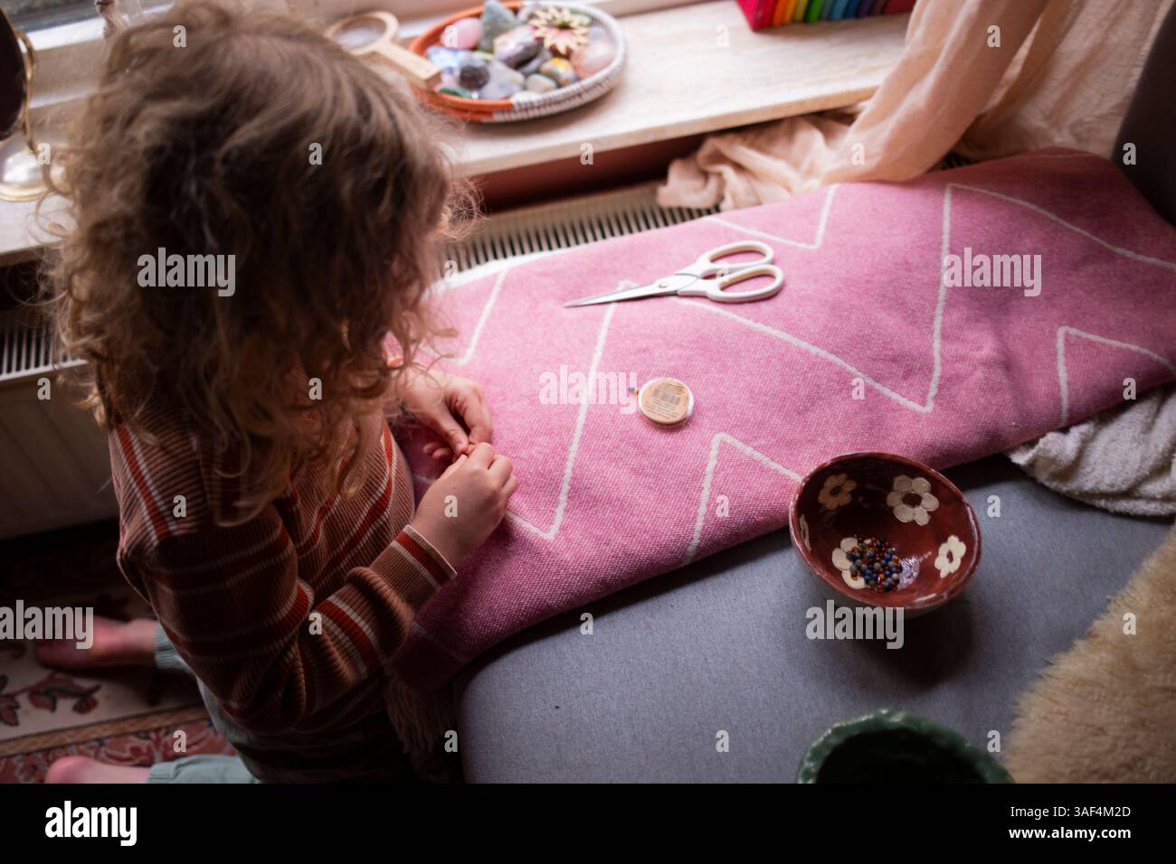 Child threading a needle while crafting on a pink textile surface Stock Photo