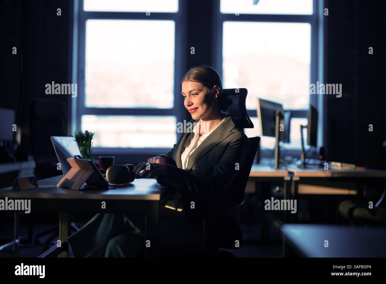 Female IT professional or AI engineer working late in a modern office, illuminated by warm sunlight. Concept of technology, software development Stock Photo