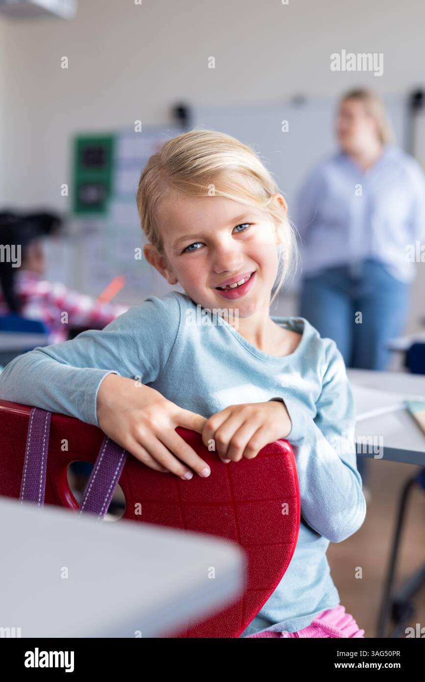 Smiling girl sitting on chair in school classroom, looking at camera ...