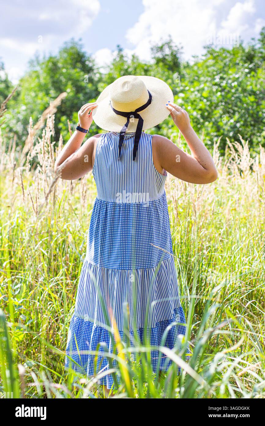 A woman wearing a straw hat stands in a field of tall grass. She is ...