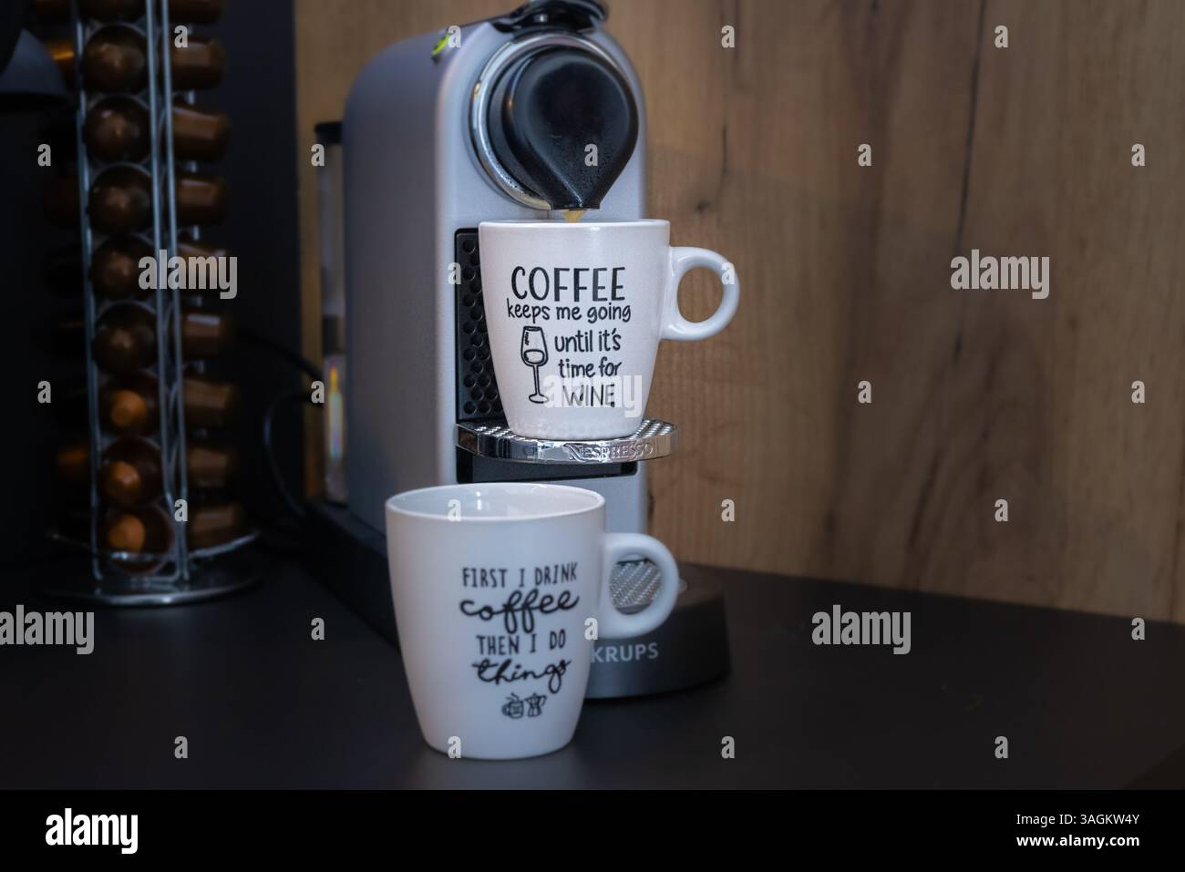 Two humorous coffee mugs sit under a Nespresso machine, one reading COFFEE keeps me going until it's time for WINE and the other first I drink coffee Stock Photo
