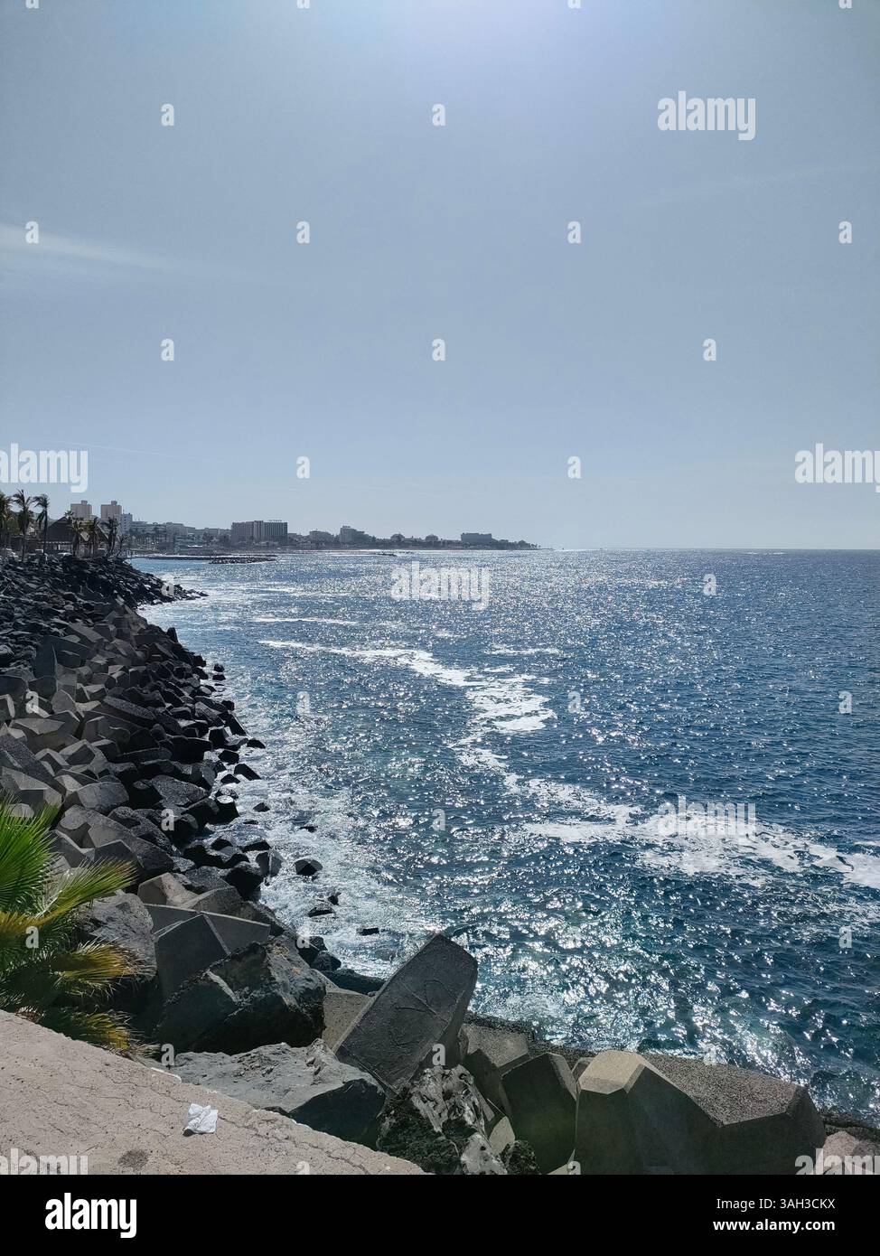 A clear coastal view with sunlight glistening on blue waves, crashing gently against a rocky shore under a wide open sky. - Smartphone Captured Stock Image