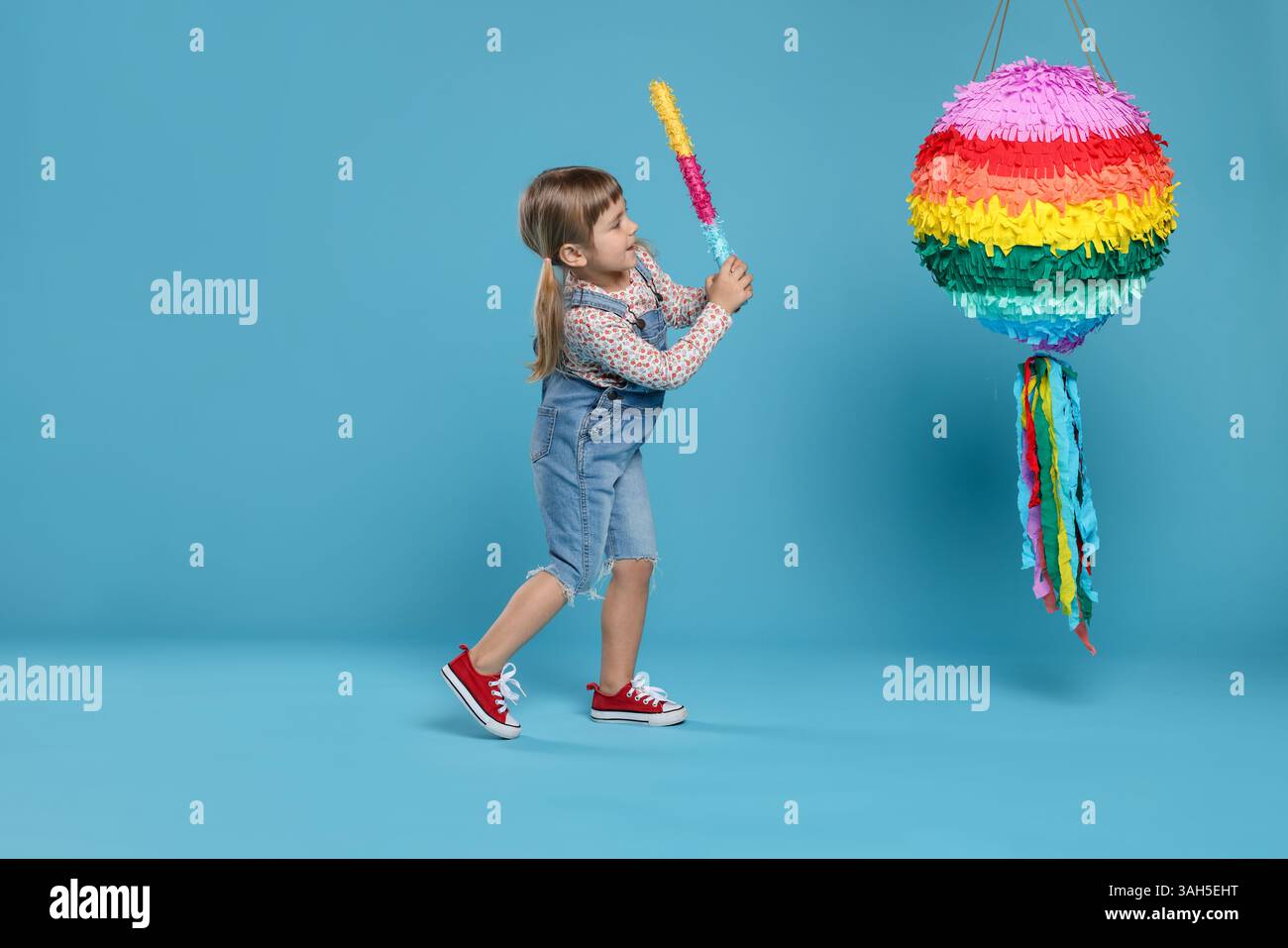 Happy girl breaking bright pinata with stick on light blue background ...