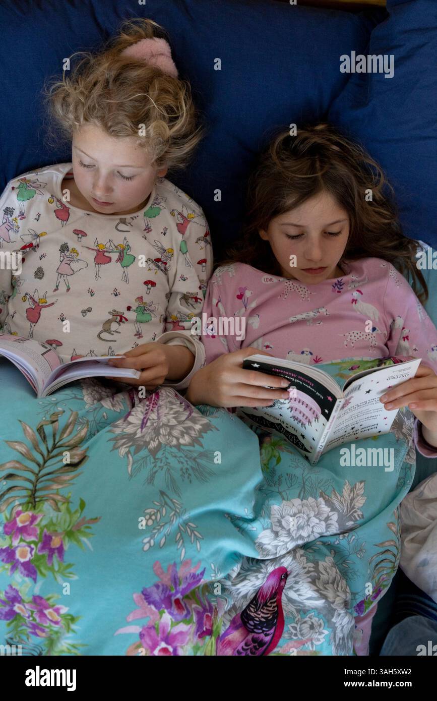 Two children sisters each reading books in bed side by side. The girls are 7 (left) and 9. Stock Photo