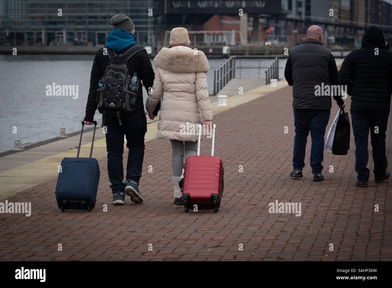 A couple strolls hand in hand along a waterfront path, pulling their luggage, as others walk in the opposite direction on a cool, day. Stock Photo