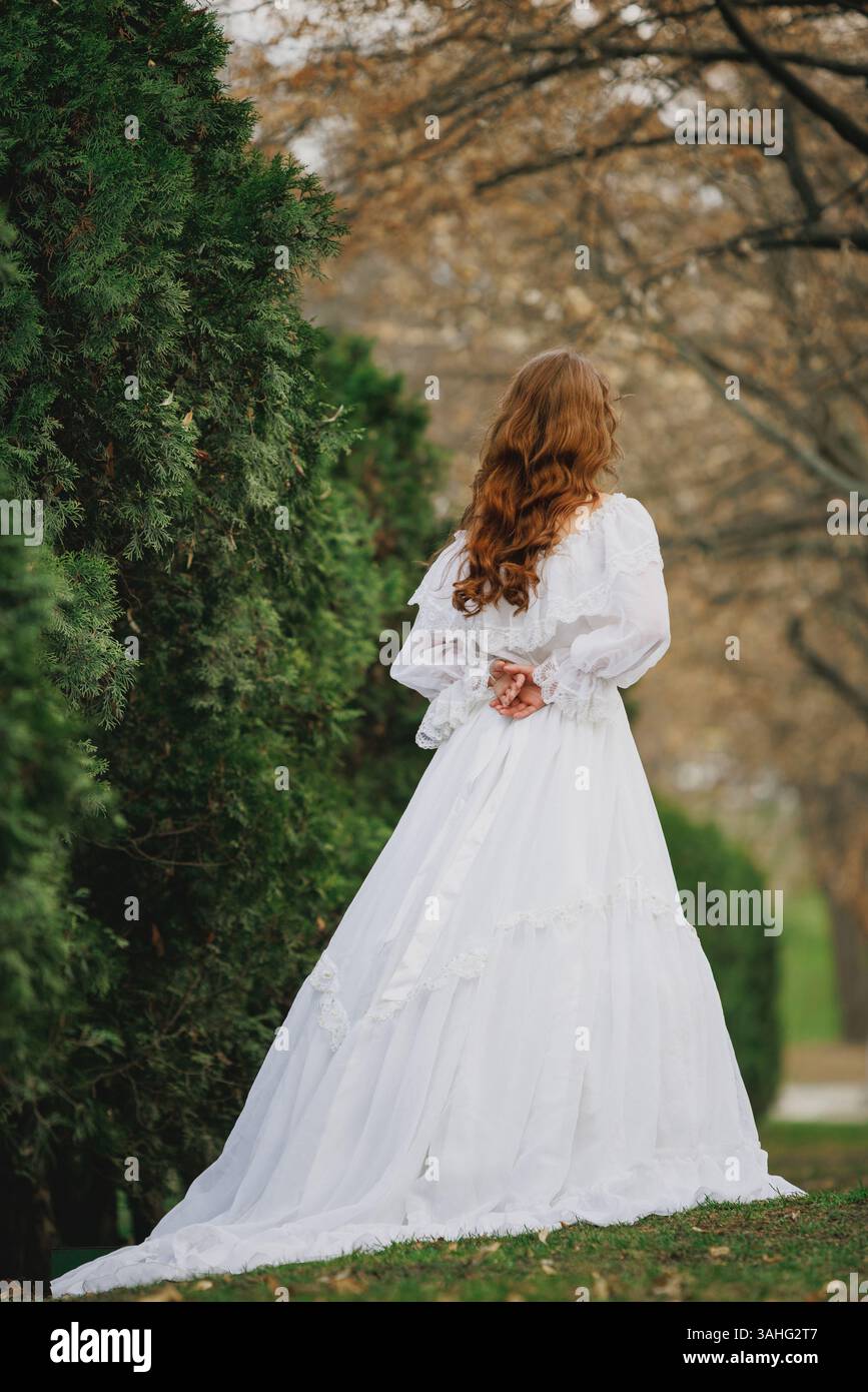 Young woman in vintage white gown walks along autumn hedge with hands folded behind back, symbolizing silence, restraint, and forgotten tales. Stock Photo