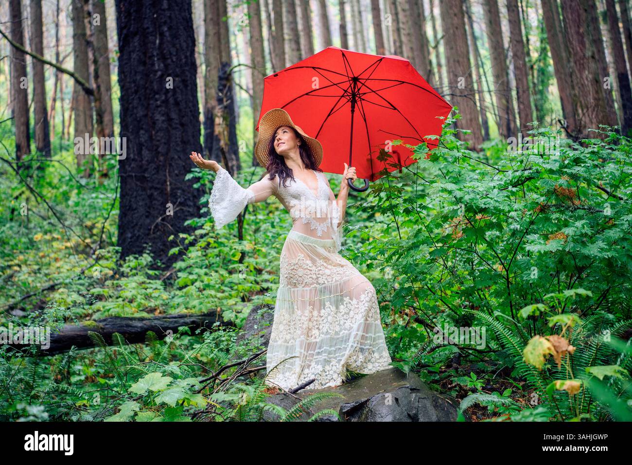 Woman holding a red umbrella in a lush, green forest wearing a white lace dress and straw hat. Silver Falls State Park, Oregon, USA Stock Photo