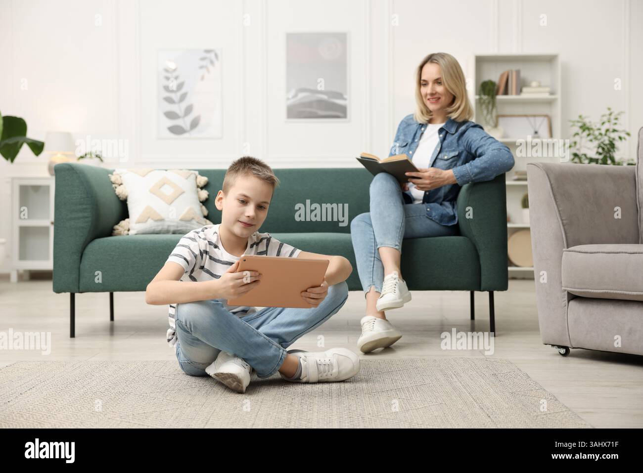 Son using tablet while his mother reading book at home, selective focus Stock Photo