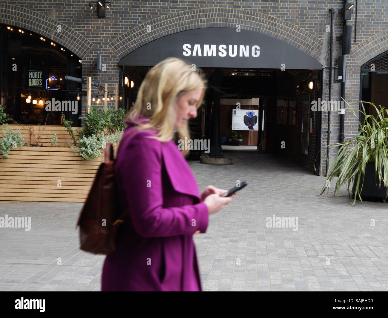 female walks past Samsung Headquarters in Kings Cross London, while looking at smartphone Stock Photo