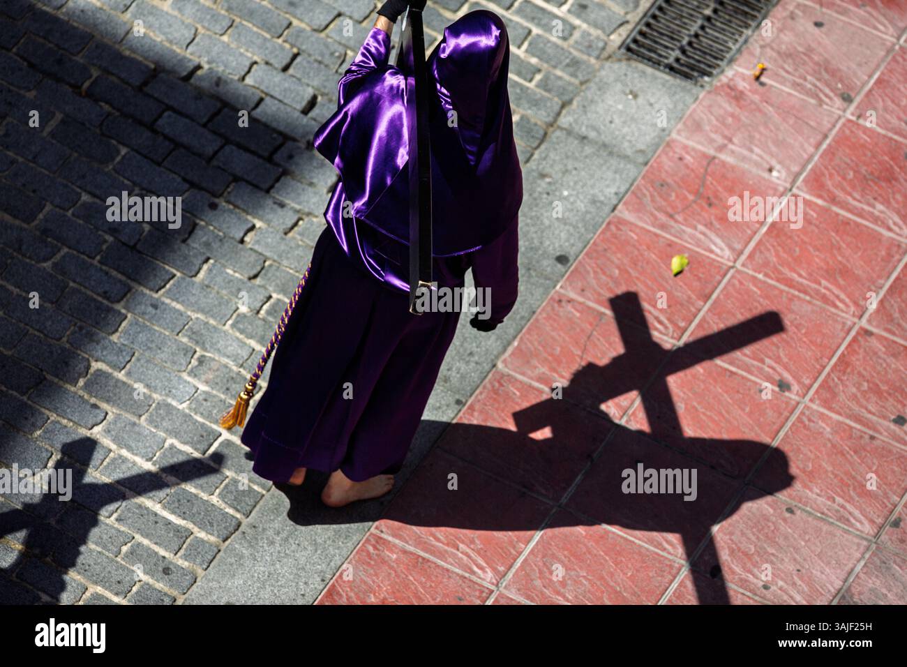 A hooded penitent walks barefoot, carrying a wooden cross through the cobbled streets of Seville during Holy Week festivities. Stock Photo