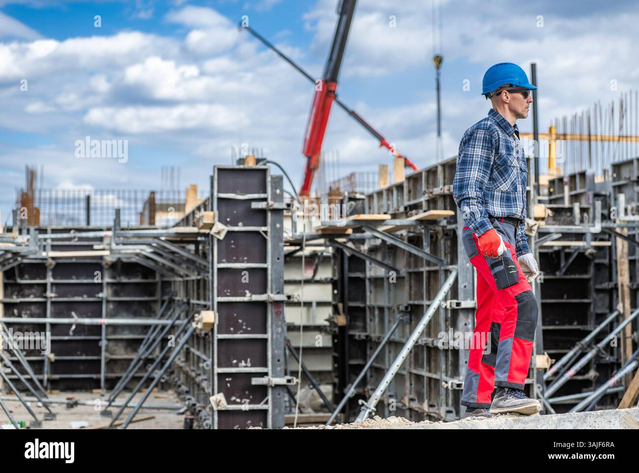 A construction worker in a helmet and checkered shirt surveys a construction site filled with scaffolding and forming systems. Cranes are visible in t Stock Photo