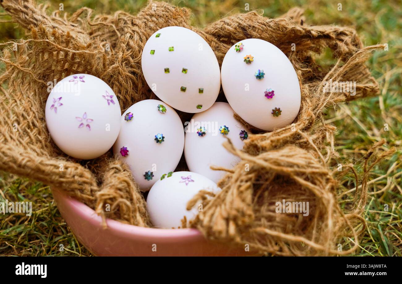 A close-up of white Easter eggs adorned with colorful sequins and arranged in a rustic jute basket on green grass, perfect for spring or Easter-themed Stock Photo